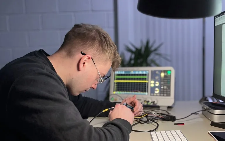 A Denotec engineer probing a circuit board on the bench, with a spectrum analyser showing a live trace behind it.