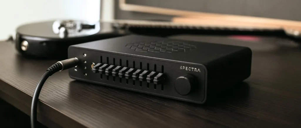 The Spectra guitar signal optimiser, a low machined aluminium unit with a row of slide controls and a rotary knob, on a dark bench with a guitar cable connected.
